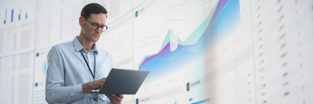 homem analisando dados em laptop ao lado de painel com gráficos